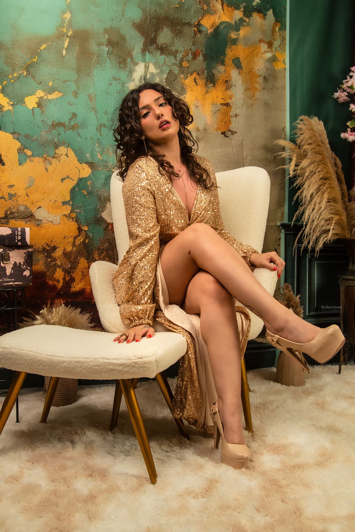 A model in a gold sequined dress seated against a teal and gold editorial backdrop.