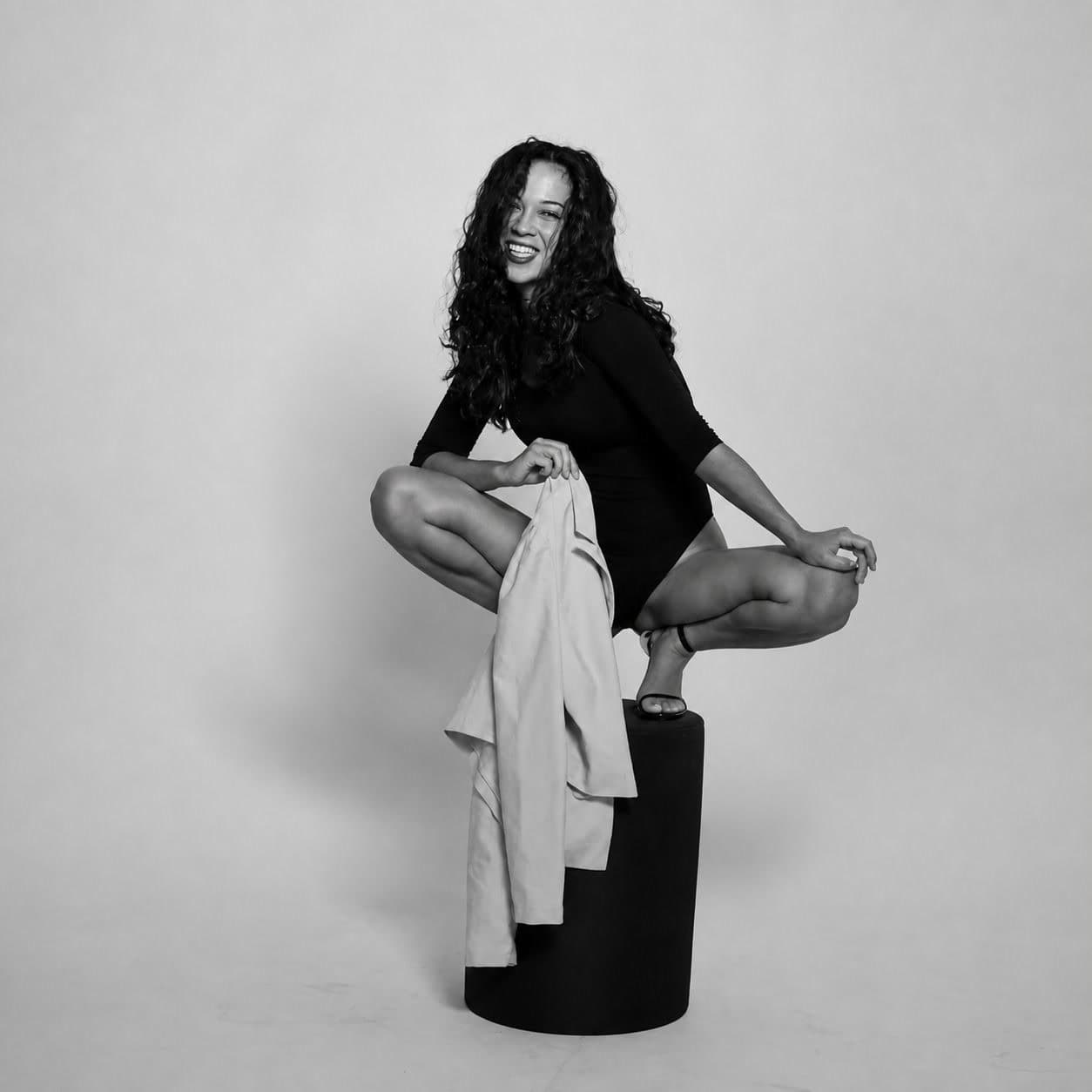 A smiling model with long curly hair posing in a black bodysuit in a black-and-white studio portrait.