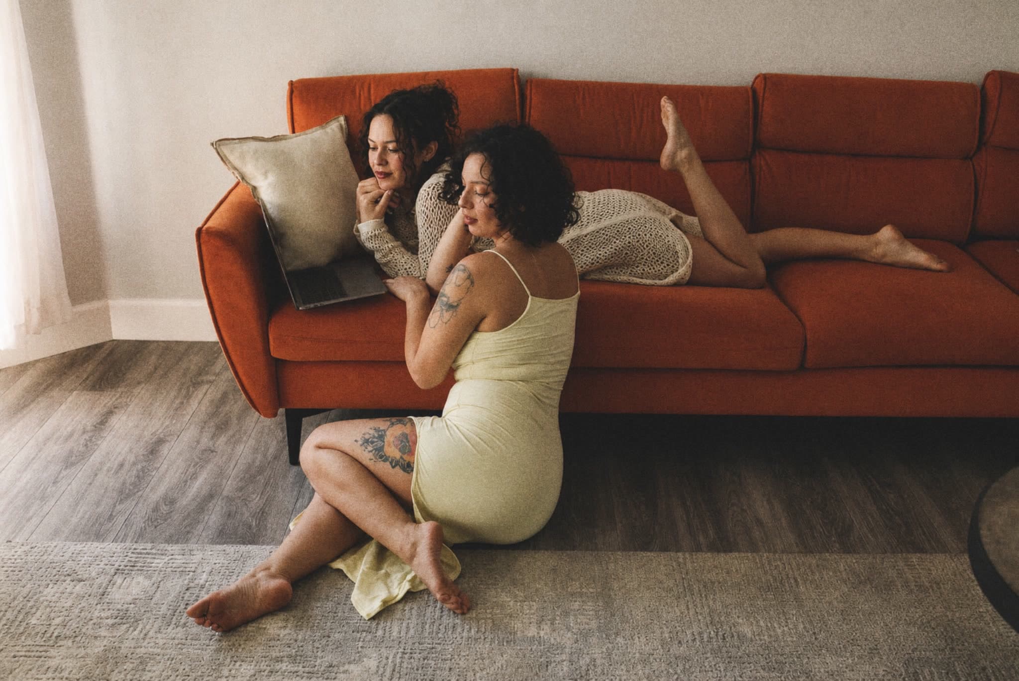 Two creative collaborators relaxing beside a laptop on a warm orange couch.